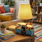 Children's room with a lamp shaped like a camper van on a table, surrounded by books and toys.
