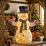 A large, festive snowman decoration made of textured white material and glowing warm white lights, with a black top hat, black-and-white check scarf, red nose, and pink-and-white mittens. It is posed in a decorated holiday home setting with a Christmas tree, wrapped gifts, and warm string lights.