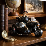 A decorative figurine of a bearded man in a red helmet riding a glossy black side-by-side motorcycle on a wooden shelf, surrounded by vintage books, a globe, and a framed picture.