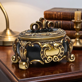 An ornate black and gold decorative box with a lid, styled on a wooden table with books and a brass lamp in the background.