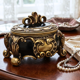 An ornate decorative lidded box with a black body and gold-toned carved detailing, placed on a wooden table with a pearl necklace nearby and a softly blurred indoor background.