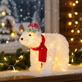 Decorative polar bear figure with a red scarf and hat on a festive background with lights and snow.