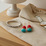 Jewelry including a silver bracelet, red and blue earrings, and a watch on a textured surface.