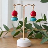 A pair of dangling earrings displayed on a gold-tone stand. Each earring has a glossy red  coral round bead on top of a glossy turquoise blue round bead, separated by a double sterling silver ring.