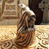 A bronze-finished figurine of a hooded, bearded Druid character sitting on a carved wooden surface, with a softly blurred decorative background.