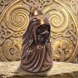 A small bronze figurine of a hooded Druid with a long nose and beard, standing on a stone surface in front of an ornate gold decorative background.