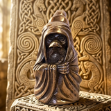 A small bronze figurine of a hooded Druid figure with a dark face and beard, posed with hands over the chest, set against an ornate carved golden background.
