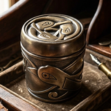 A small cylindrical metal container with a lid, decorated with embossed bird and scroll motifs, shown in a rustic wooden tray next to a brass pen nib.