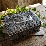 A rectangular metal keepsake box with embossed Celtic knotwork patterns and a central relief panel, displayed on a rustic wooden surface with green foliage around it.