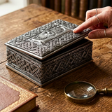 An ornate rectangular metal box with a lid, featuring embossed Celtic knot patterns and a central relief of a Greenman, shown on a wooden table with a hand lifting the lid. A magnifying glass and a book are nearby.
