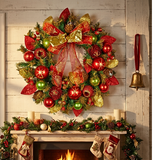 A large festive Christmas wreath with red and gold ribbons, red and green ornaments, pinecones, and gold star accents, hanging above a decorated fireplace mantel with stockings and a lit fire. A gold bell hangs on the wall to the right.