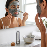 Woman applying face mask in front of a mirror with a product on the counter.