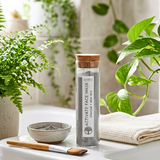 Glass jar with cork lid containing an activate face mask, surrounded by plants on a white surface.