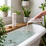 Person pouring a bath product into a bathtub with plants around