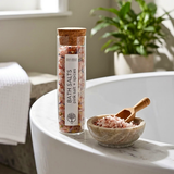 A clear cylindrical bottle of bath salts labeled  BATH SALTS PINK SALT + SPICES' with a cork stopper, standing on a white bathroom ledge beside a small bowl of pink bath salts and a wooden scoop.