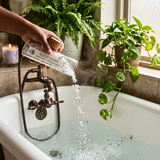 A hand holds a clear bottle labeled 'SLEEP SOAK' and pours white granules into a white bathtub with warm water and an antique-style faucet. Green potted plants and a lit candle sit on the windowsill in a bright bathroom setting.