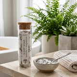 A glass jar labeled 'SLEEP SOAK lavender & magnesium' with a cork lid stands on a stone surface beside a small bowl of the soak and a spoon, with a folded towel nearby and potted plants in the background.