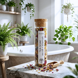 A clear cylindrical jar with a cork lid and white label reading 'TEA SOAK sea salt + botanicals' stands on a marble surface in a bright bathroom. The jar contains a colorful loose-leaf tea blend with visible sea salt and dried flowers.