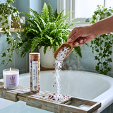 A hand pours white bath salts from a wooden scoop into a bathtub, with a jar labeled 'Tea Soak' and a lit lavender candle on a wooden bath caddy. The scene is styled in a bright bathroom with green plants and a window.