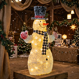 A large illuminated snowman decoration standing on a wooden platform in a rustic indoor holiday setting. The snowman has a textured white body with warm yellow lights, a black top hat with red trim, a black-and-white plaid scarf, red mittens, and a carrot nose.