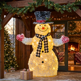 A large illuminated snowman holiday decoration stands on a stone patio in front of a wooden porch with a lit fireplace and Christmas tree in the background. The snowman has a glowing yellow body, a black-and-white check scarf, red-and-white mittens, and a black top hat with a red band.