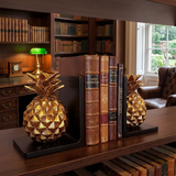 Decorative pineapple bookends on a wooden surface with books and a lamp in the background.