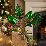 Green glittery acorns with leaf-shaped tops and jeweled bands, attached to twine and displayed among pine branches and burlap in a holiday setting.