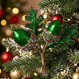 A close-up of a green acorn Christmas ornament with glittery green leaves and a brown branch, hanging on a decorated Christmas tree with red and gold ornaments and warm string lights.