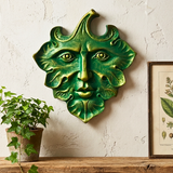 Green wall art with face design on a textured wall with a plant and framed picture.