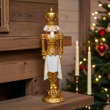Decorative nutcracker figure in gold and white on a wooden surface with a Christmas tree in the background.