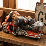 Decorative figurine of a man lying on a motorcycle with a suitcase on a wooden surface.