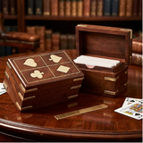 A wooden card deck box set shown on a polished wooden table with a blurred library background. One closed box displays four engraved suit symbols (hearts, clubs, diamonds, and a square) on the lid, while another open box reveals a stack of white playing cards.