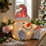 A festive snowman-shaped holiday light decoration with a red Santa hat, glowing warm lights, an orange carrot nose, and gray snow-covered mittens, displayed on a wooden table with Christmas greenery and a lit tree in the background.
