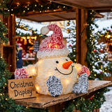 A festive Christmas snowman decoration made from textured white material, wearing a red Santa hat with white snow-like trim, an orange carrot nose, and gray-and-white mittens, displayed on a wooden table at a holiday market with warm string lights and green garlands in the background.
