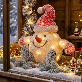 A festive snowman holiday decoration with warm white lights, a red Santa hat, and red pom-poms, displayed on a wooden shelf with Christmas trees and lights in the background.