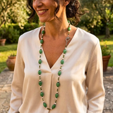 A woman wearing a long gemstone necklace with green oval stones and a sterling silver clasp, paired with a cream blouse, photographed outdoors in a garden setting.
