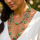 A close-up of a woman wearing a layered necklace with green adventurine stone beads, small white pearls beads, and a sterling silver bird clasp.