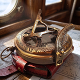 A brass and bronze vintage-style compass with an attached leather case, photographed on a wooden surface near a window with warm sunlight and dust particles visible.