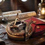 A brass pocket compass with a red leather case sits on a wooden table, surrounded by vintage props including a lantern, ink bottle, and rolled map in a warm, atmospheric setting.