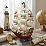 A decorative metal and resin sailing ship model with cream sails and a red hull sits on a rustic table, surrounded by nautical props including a lighthouse, globe, compass, and scrolls in a softly lit indoor setting.