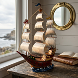 A decorative metal and resin sailing ship model with cream sails and a reddish-brown hull sits on a rustic wooden table beside an open book and a brass porthole mirror, with a window showing a coastal view in the background.