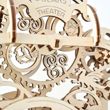 Close-up of a wooden mechanical puzzle or decorative sculpture with interlocking laser-cut gears, rings, and fantasy creature silhouettes, including a dragon and a face cutout. The words 'AGE' and 'THEATER' are visible on the wood.