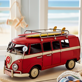 Red model van with a surfboard on top, placed indoors near a window with a view of the ocean.