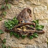 A whimsical wall-mounted sculpture of a goblin or fairy face emerging from green ivy leaves, wearing a pointed brown hat with a small gold buckle. The piece is mounted on a textured stone wall with climbing ivy.